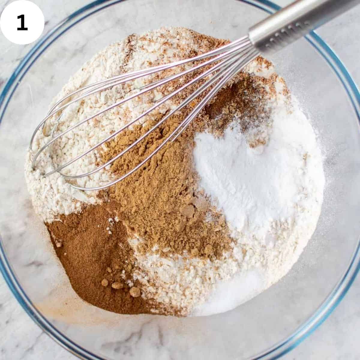 Different colored dry ingredients in a glass bowl with a whisk.