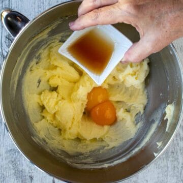 Creamy yellow mixture in the bottom of a steel bowl along with two eggs and vanilla extract being poured in.