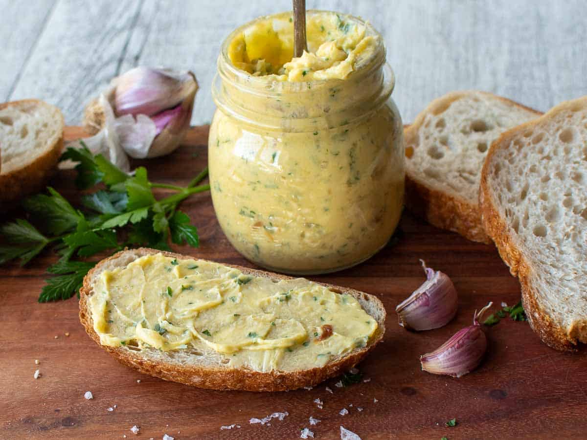 Softened butter with green herbs in a jar and also spread onto bread on a wooden board.