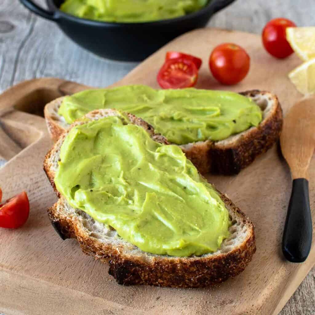 Avocado Spread on two slices of sourdough bread, cherry tomatoes and more spread in background.