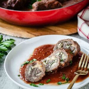 An Authentic Italian Braciole recipe prepare in a red pan with one braciole sliced on a white plate.