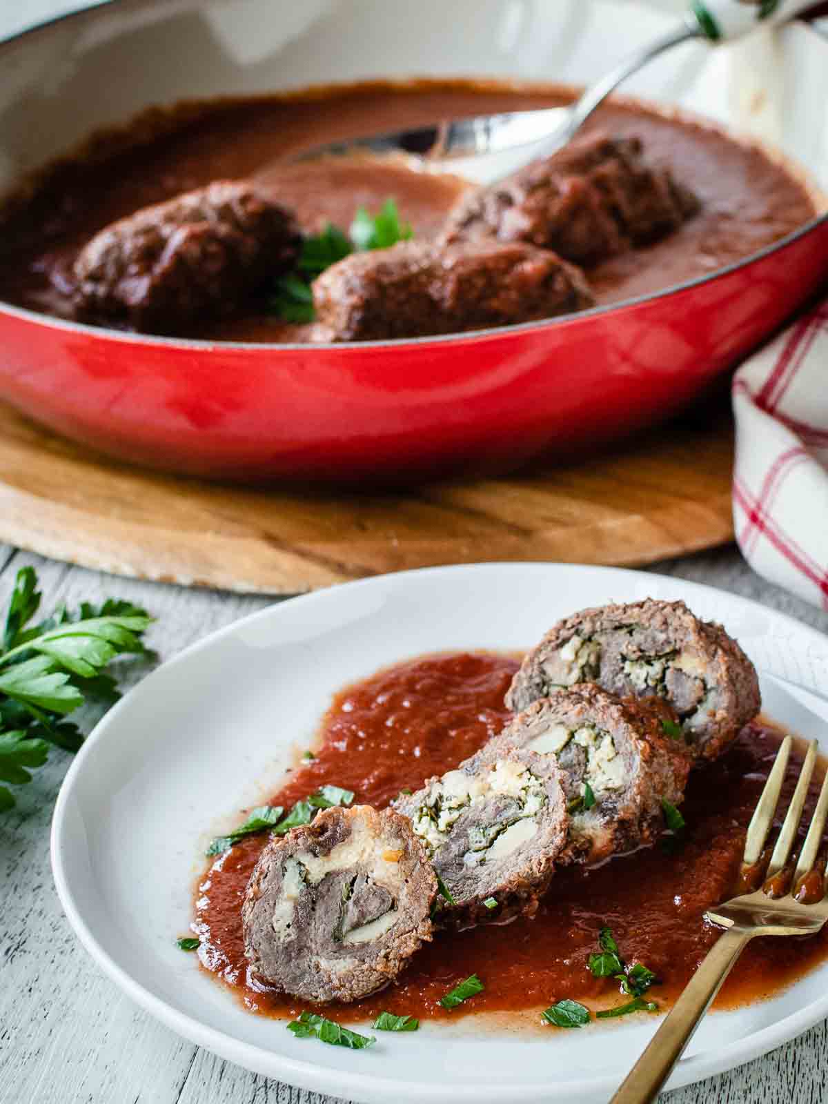 An Authentic Italian Braciole recipe prepare in a red pan with one braciole sliced on a white plate.