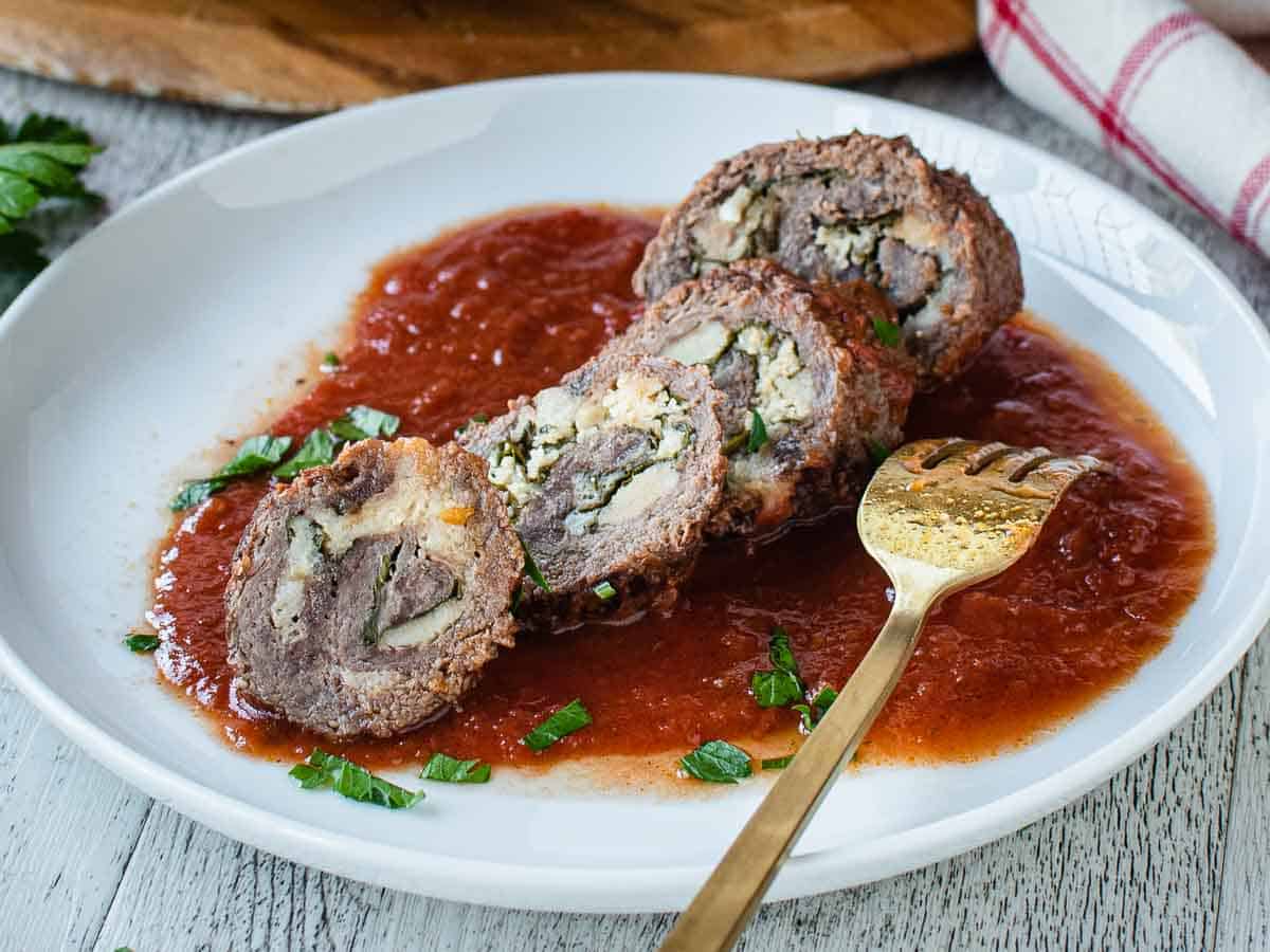 Beef braciole sliced and served on tomato sauce on a white plate.