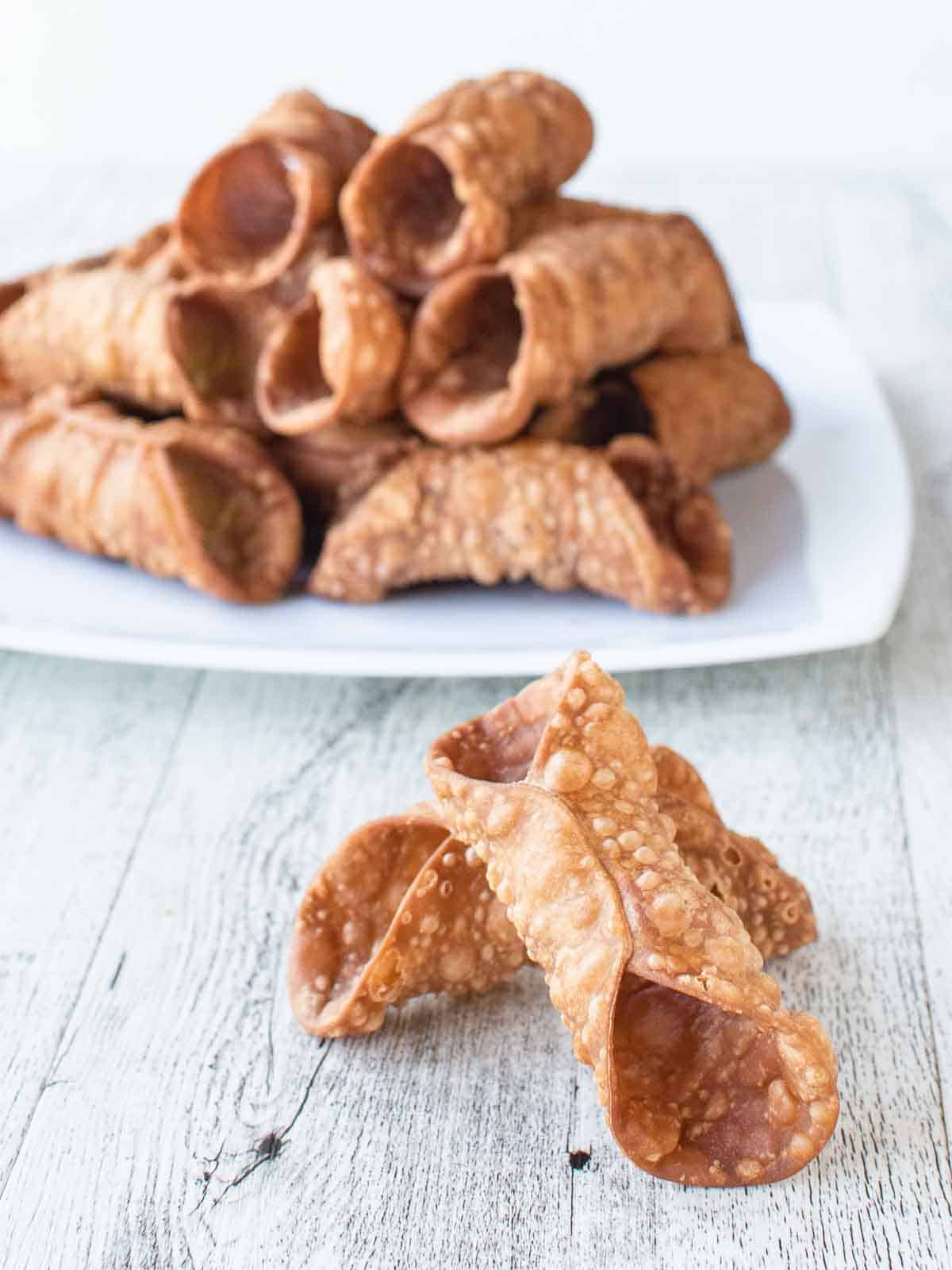 Two Cannoli Shells in front of a white plate piled high with cannoli shells.