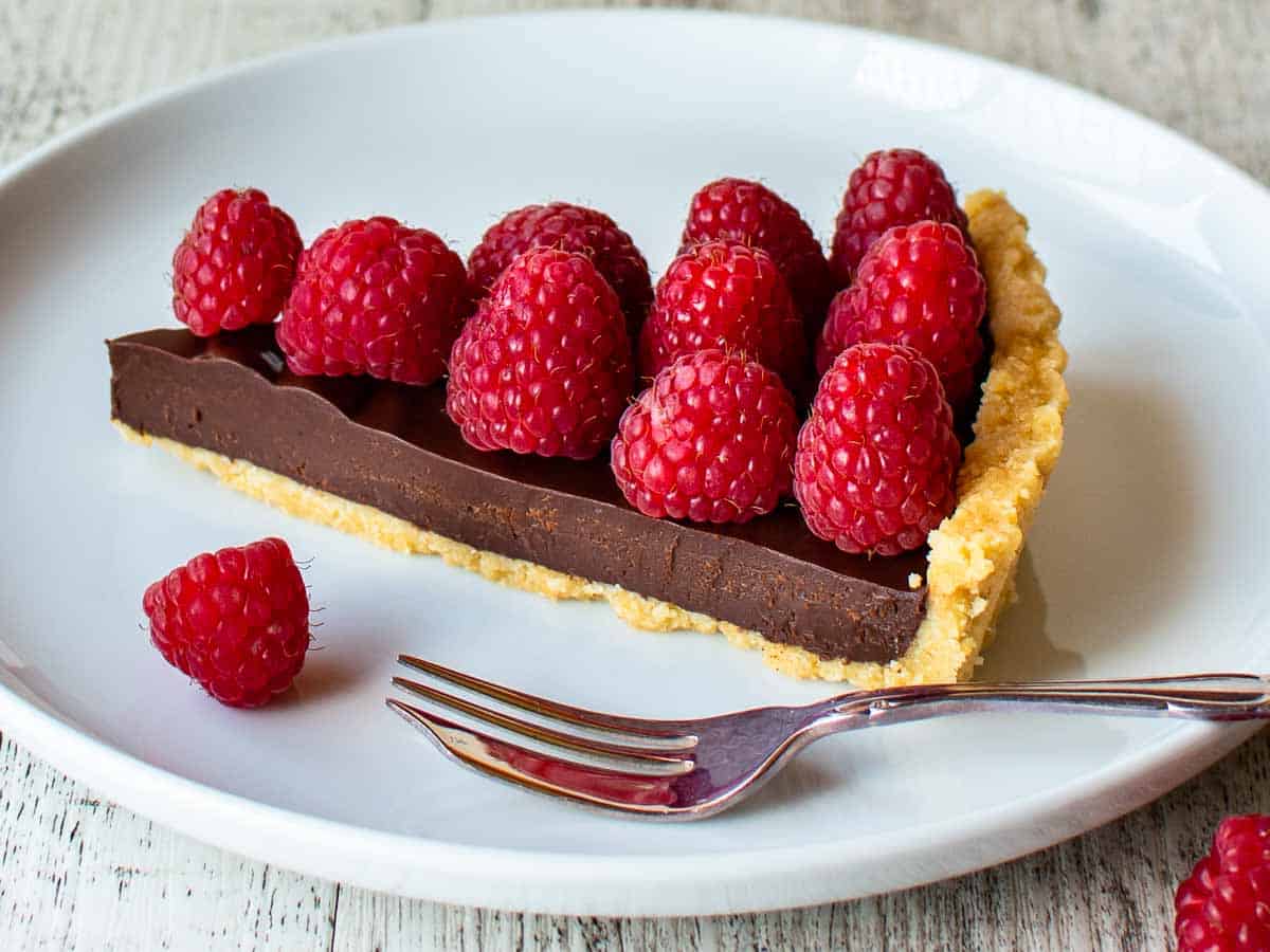 Wedge of chocolate tart with raspberries on top on a white plate.