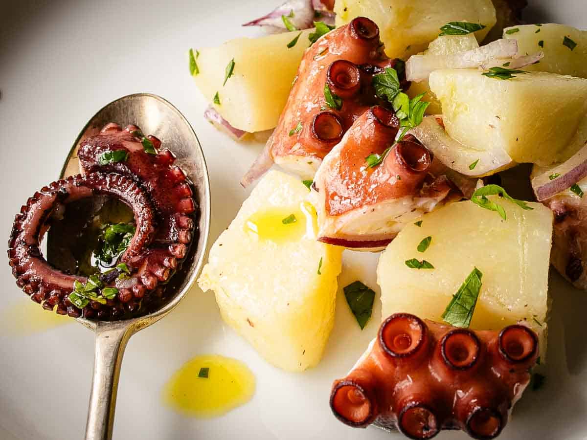 The thin end of octopus tentacle rolled up in a spoon with potato cubes and pieces of octopus tentacles beside.
