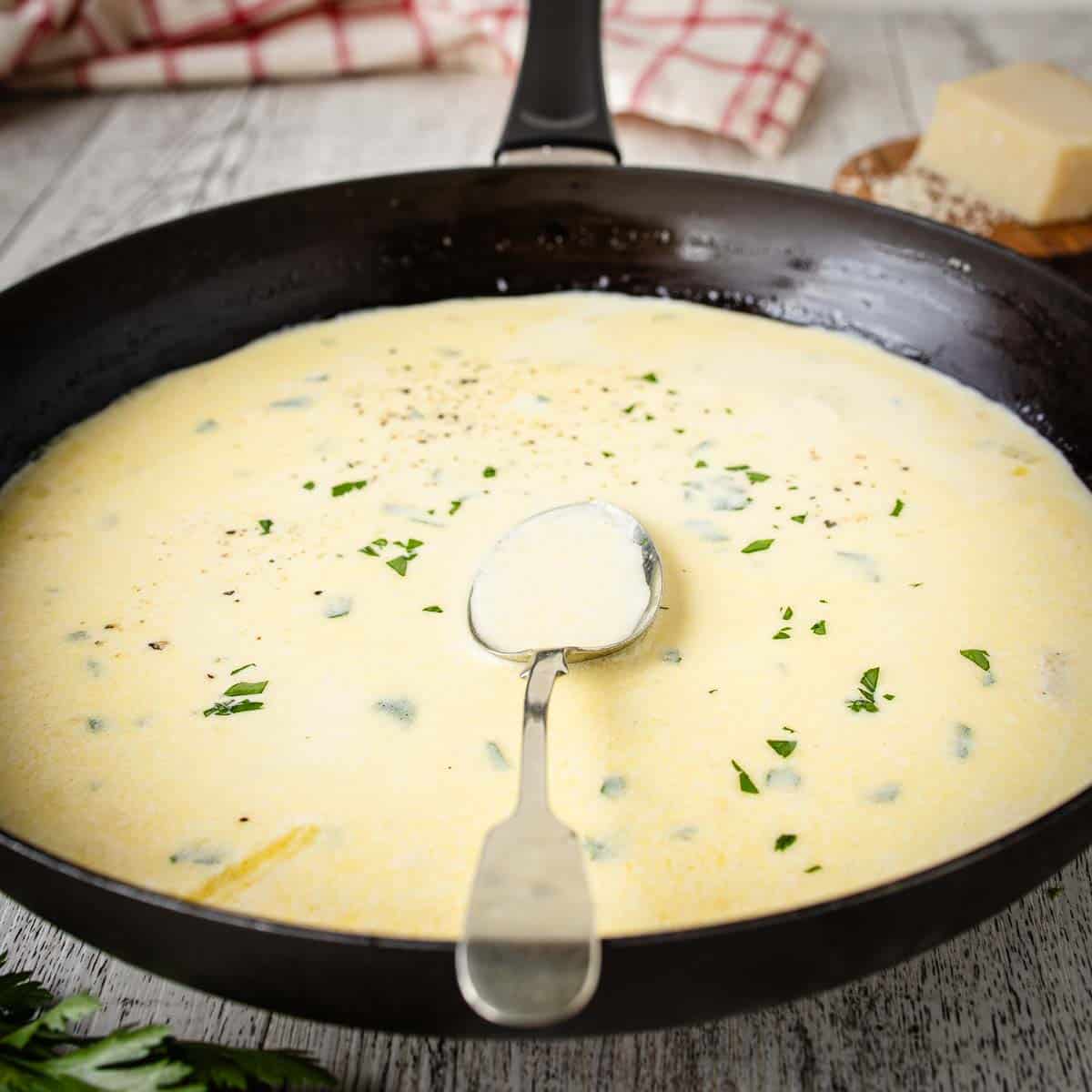 A close-up of a skillet filled with Garlic Parmesan sauce, with a metal spoon resting in it.