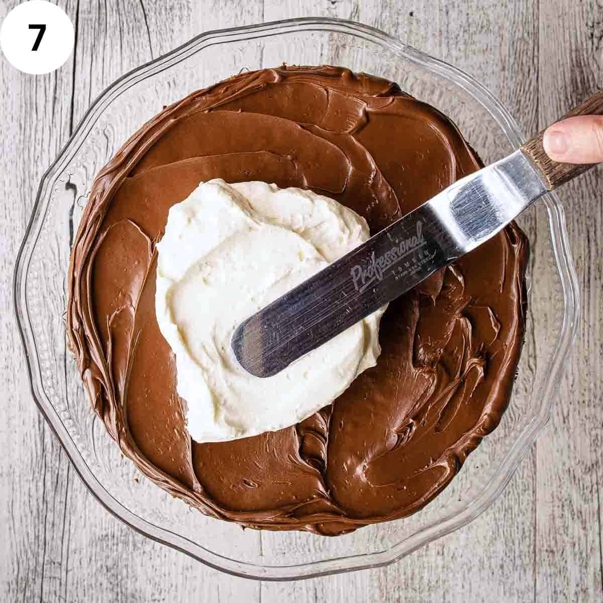 Whipped cream being spread onto chocolate cake topped with chocolate spread.