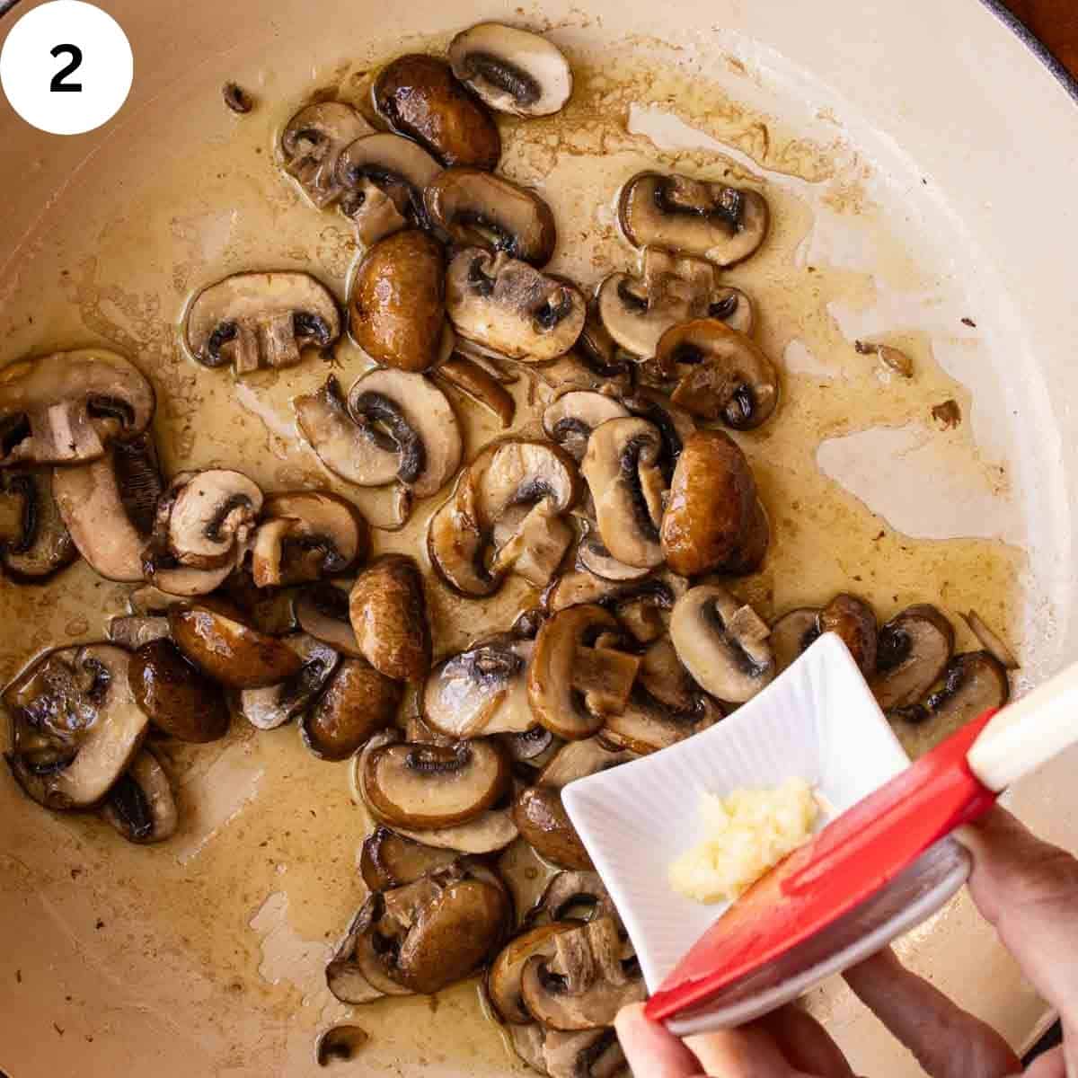 Cooked sliced mushrooms in a skillet with minced garlic being added.