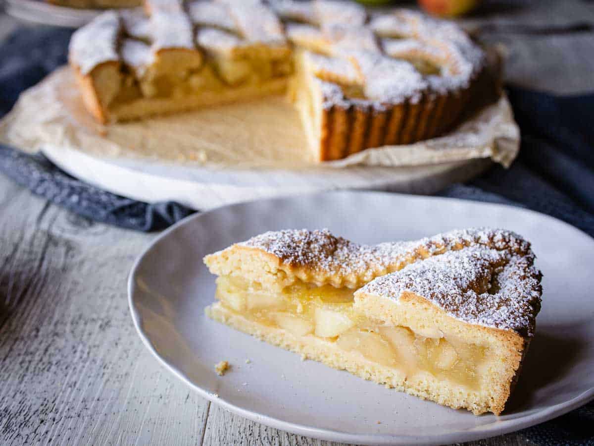 Wedge of tart filled with apple on a plate with whole tart in the background.