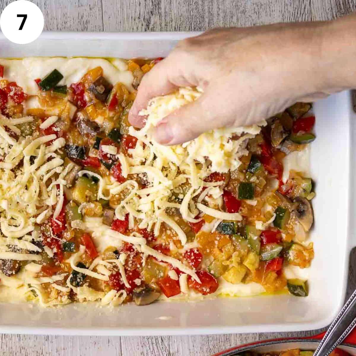 Sprinkling cheese over cooked mixed  vegetables in a lasagna dish.