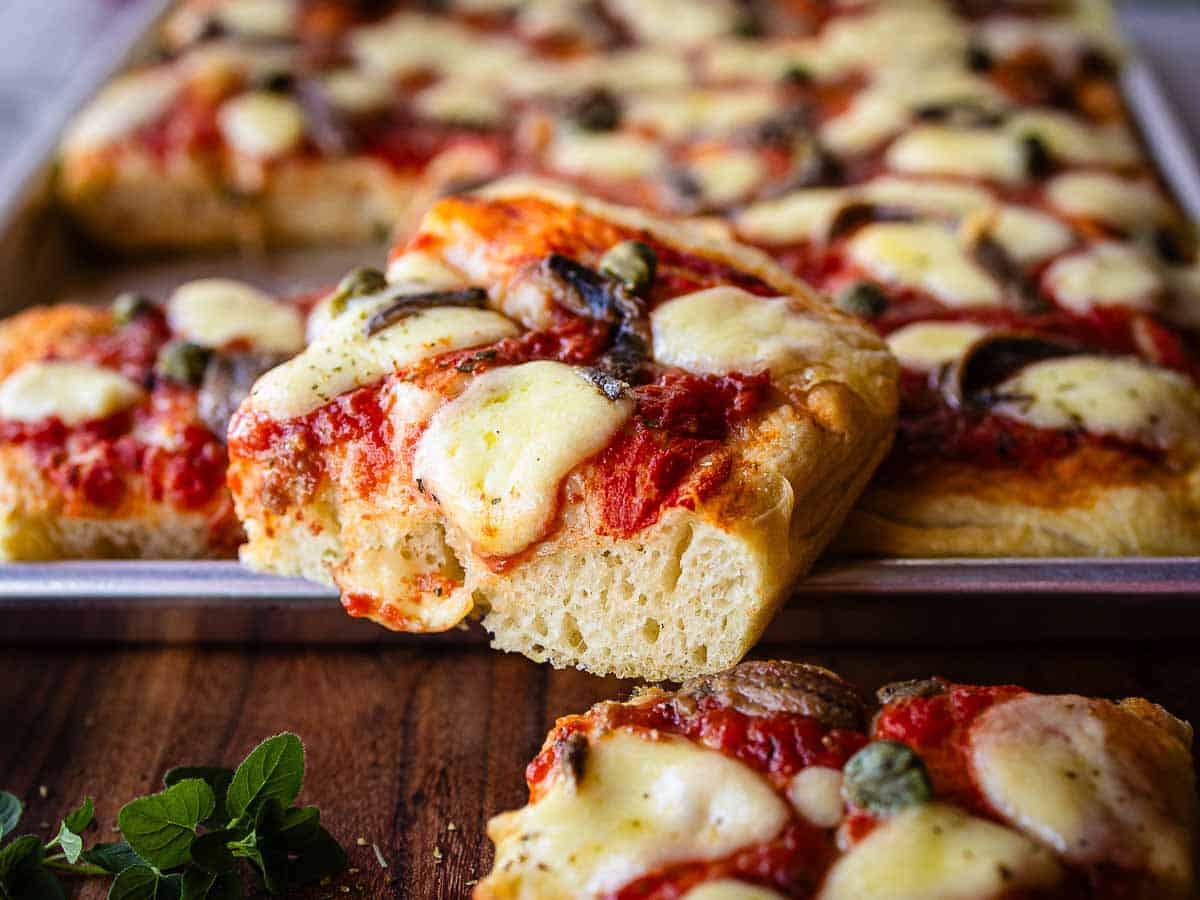 A square slice of thick crusted pizza resting on the edge of the rectangular pizza pan.