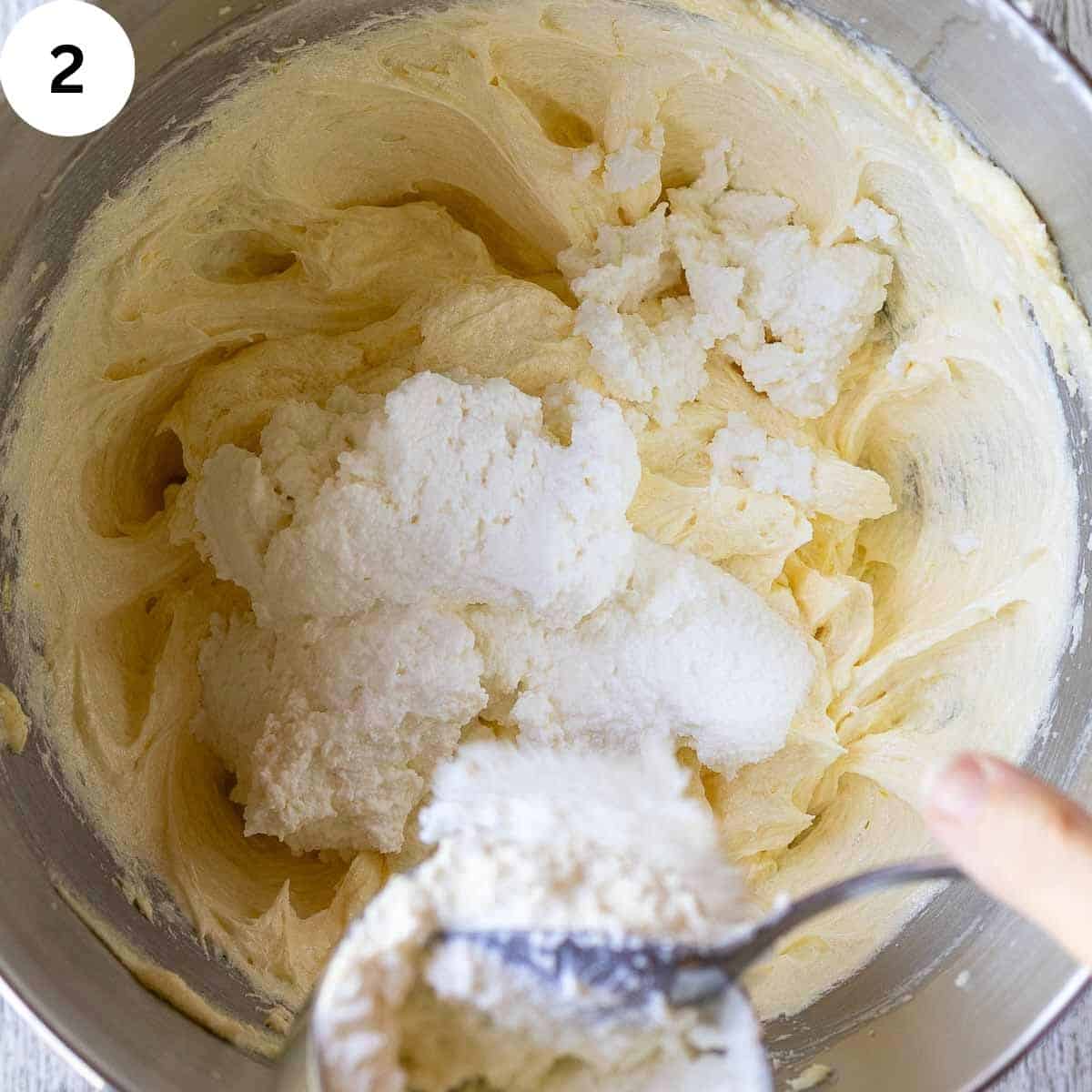 Ricotta cheese being scraped onto a creamy butter and sugar mixture.
