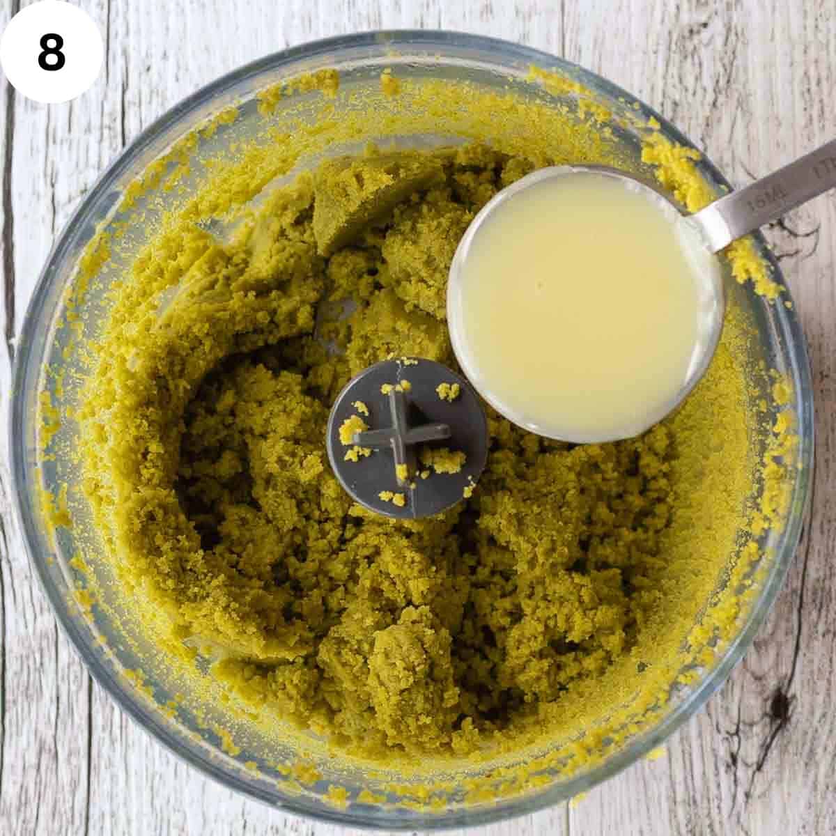 A spoonful of creamy liquid being poured into food processor containing ground pistachios.