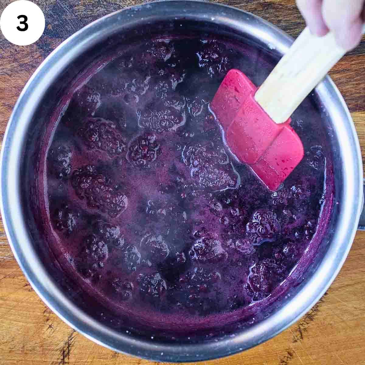 Blackberries simmering in a saucepan being stirred with a spatula.
