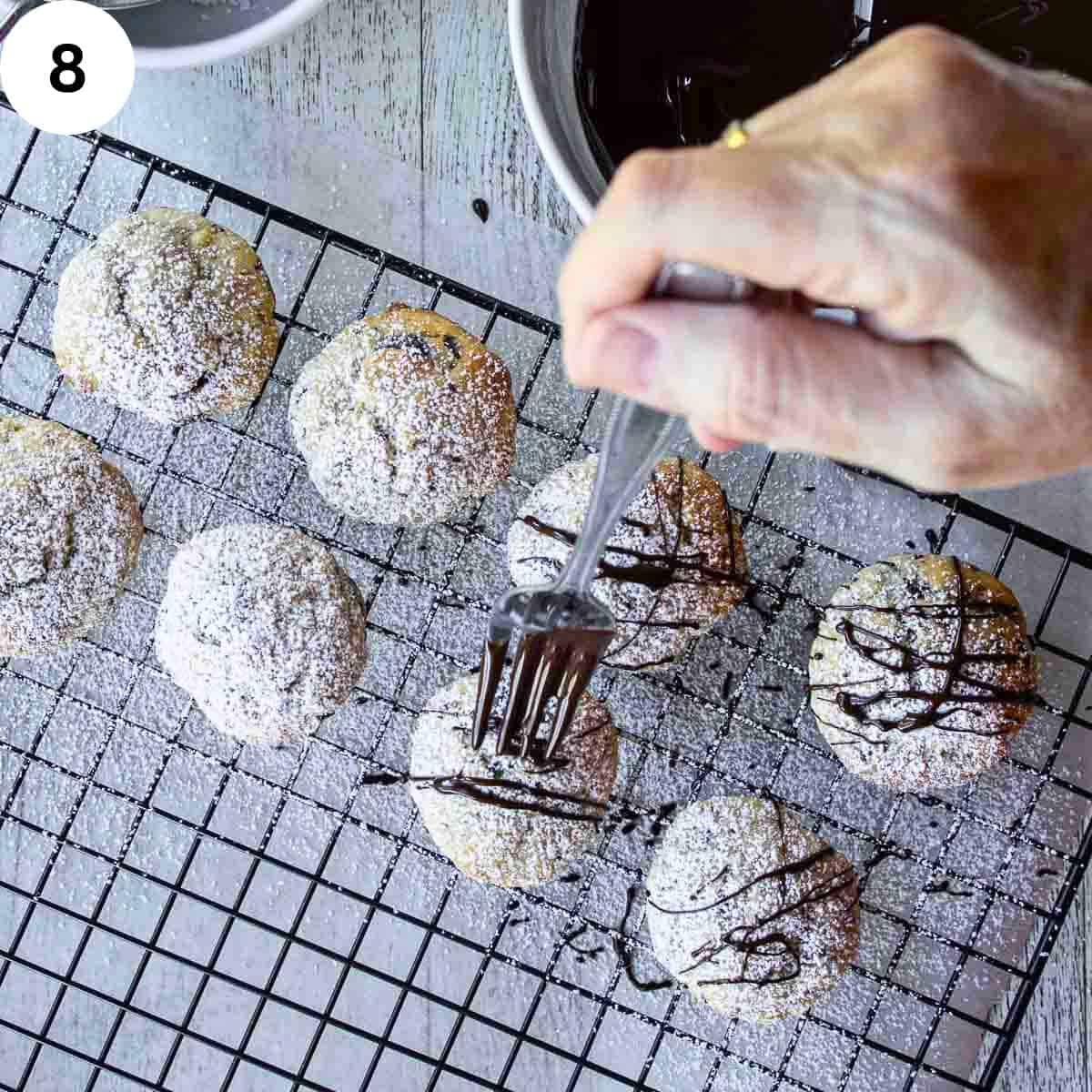 A person drizzling melted chocolate using a fork over a tray of cookies.