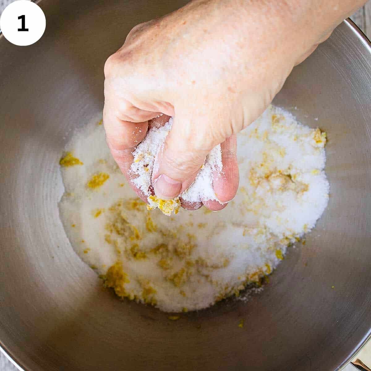 A hand is stirring a mixture of orange zest and sugar in a bowl.