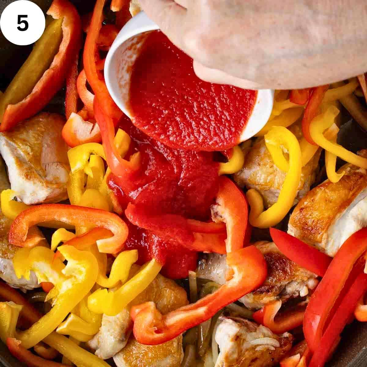 A person adding tomato passata to a pan of fried chicken and sliced bell peppers.