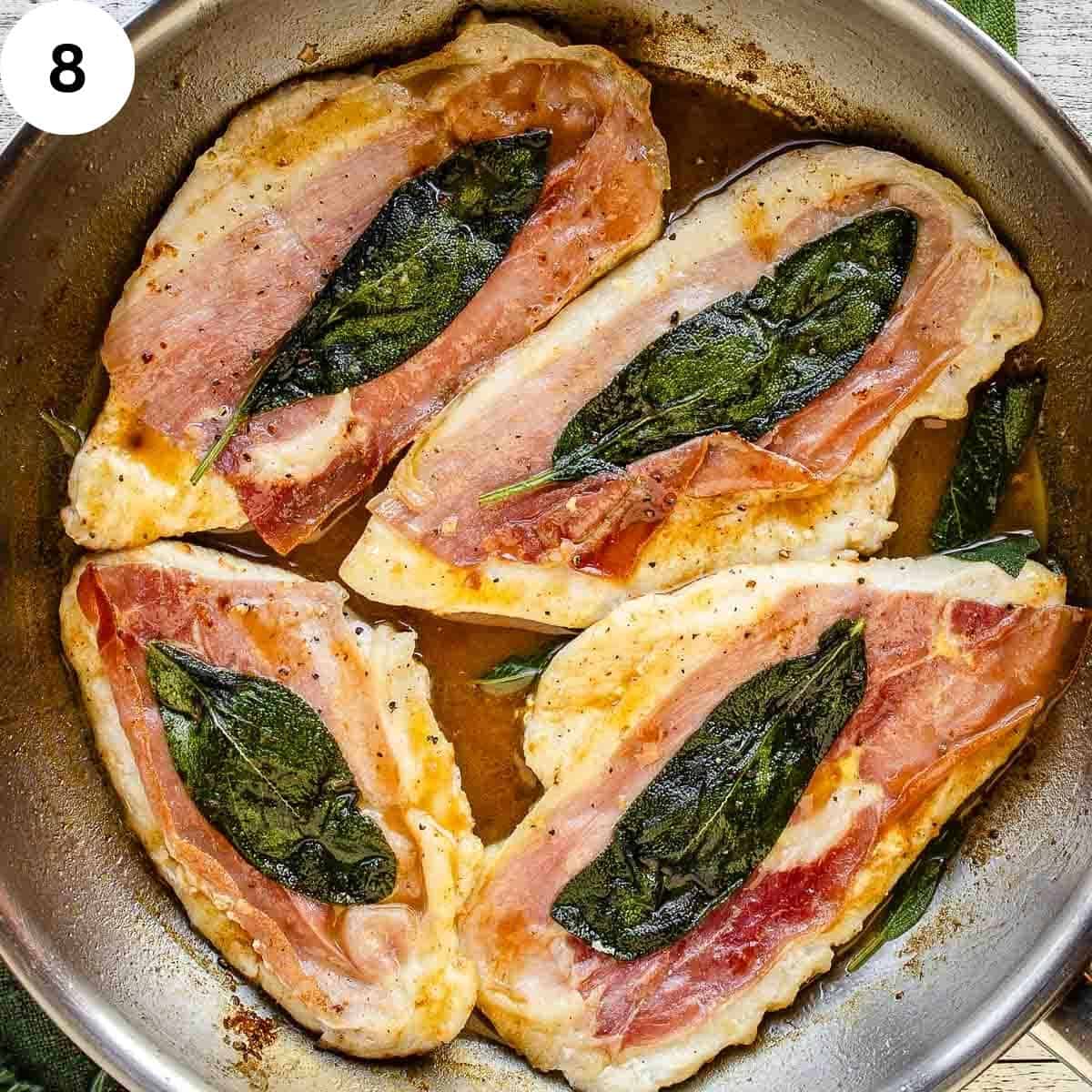 Four cooked chicken breasts each with prosciutto and a sage leaf in a pan.