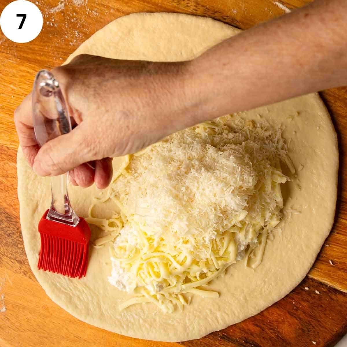 Brushing water along the edge of rolled dough before sealing a cheese filled calzone.