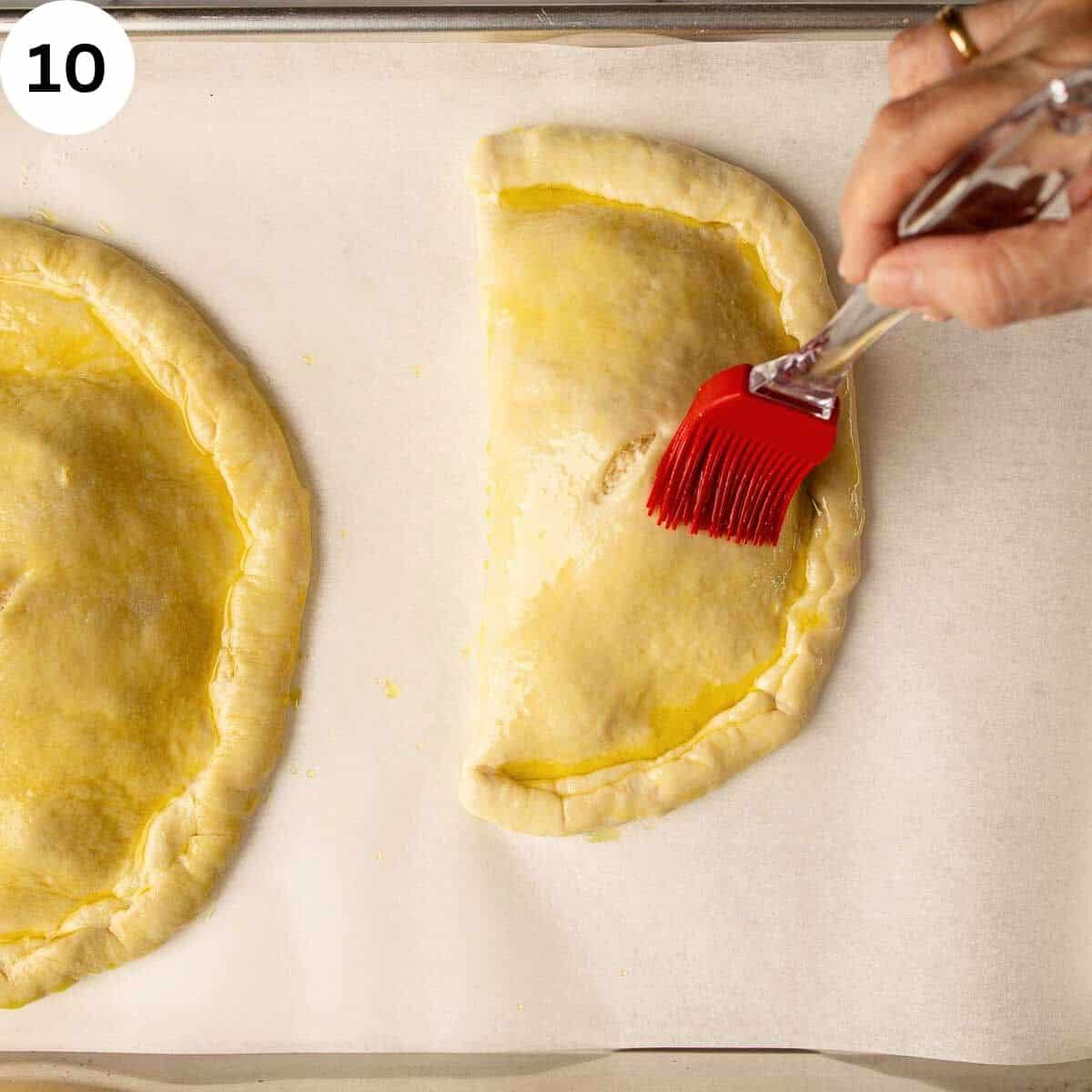 Brushing oil over shaped calzones on a lined baking tray before baking.