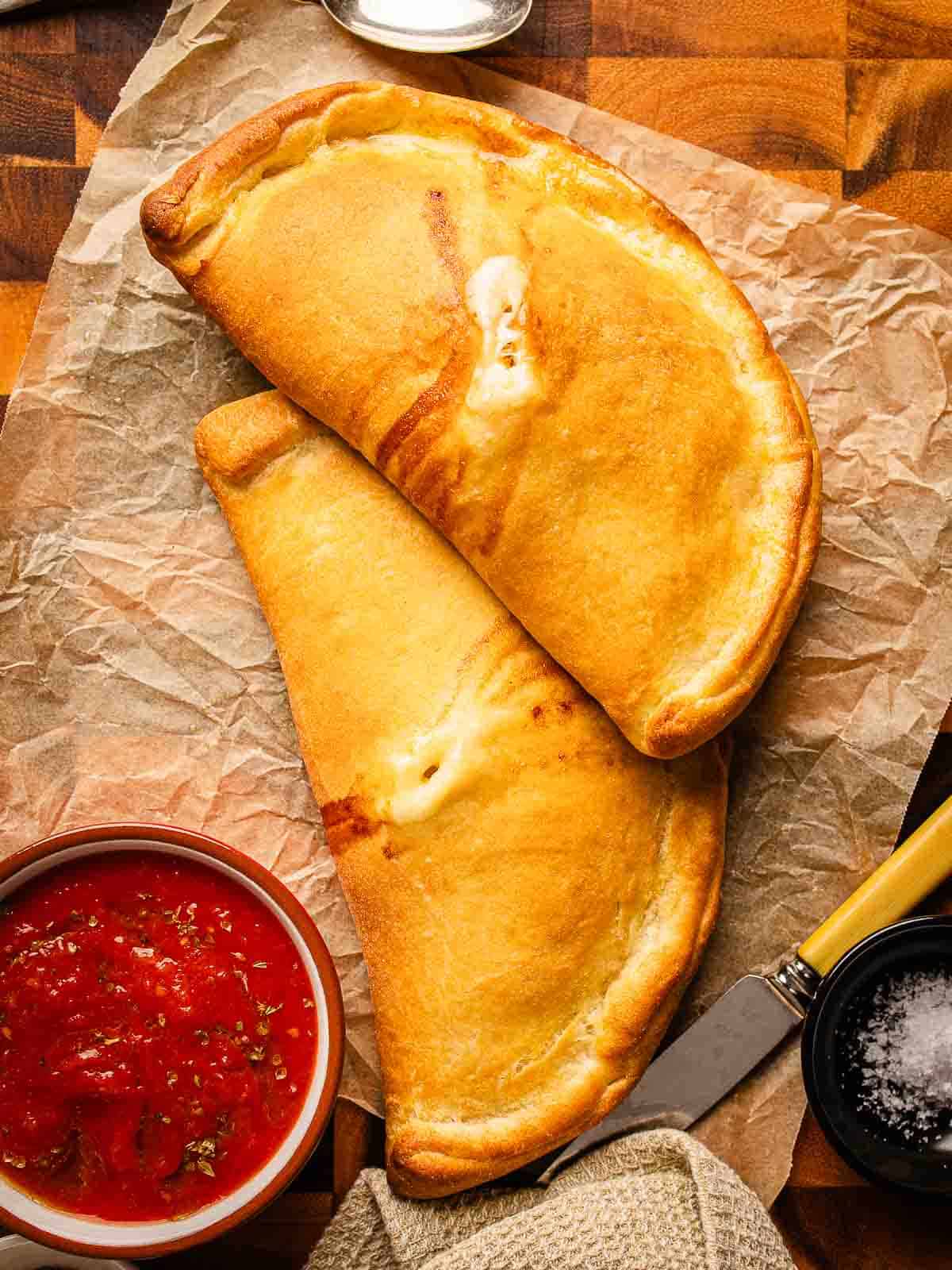 Overhead view of two baked calzone with marinara sauce in a bowl nearby.