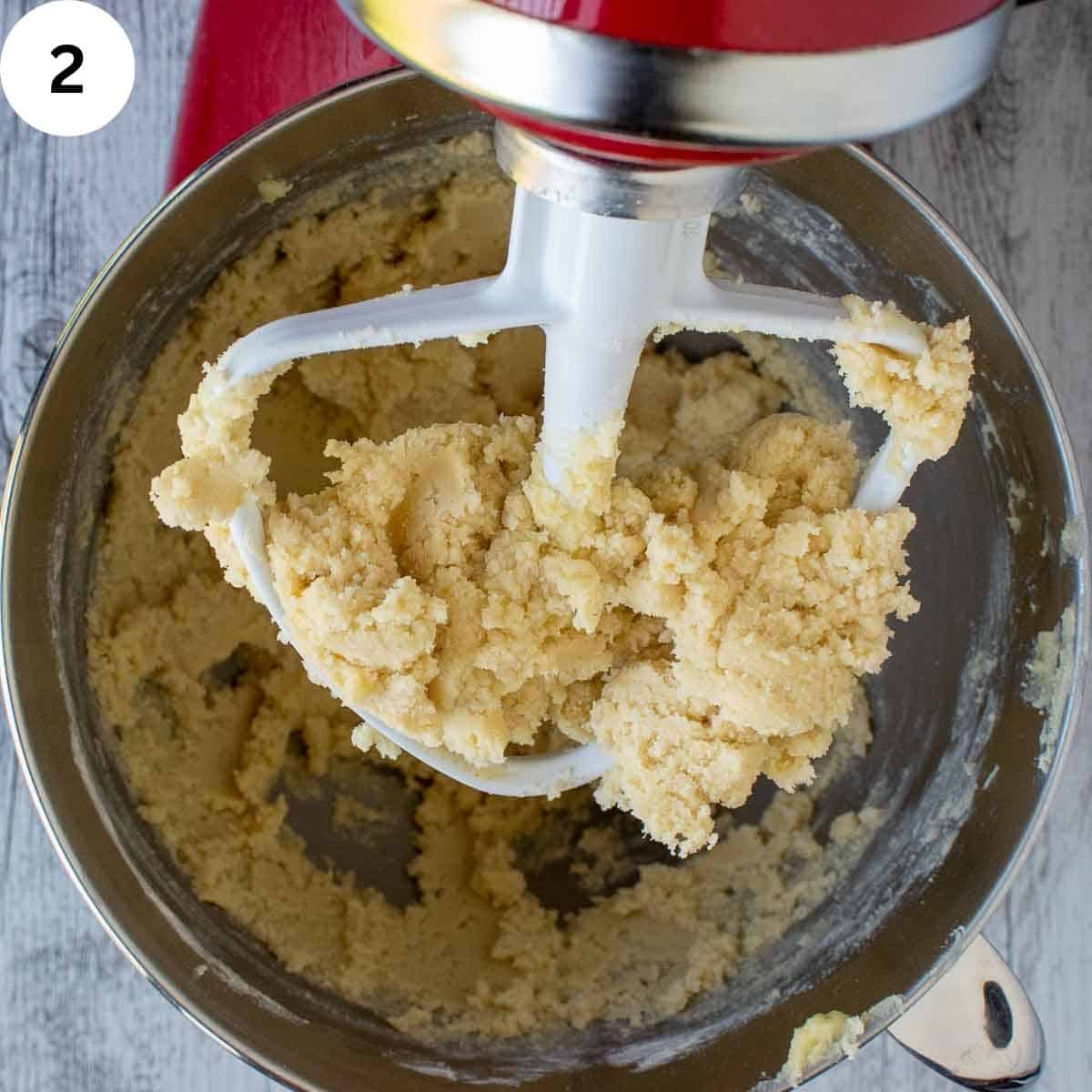 The paddle of an electric mixer covered with cookie dough suspended above a bowl of the same cookie dough.