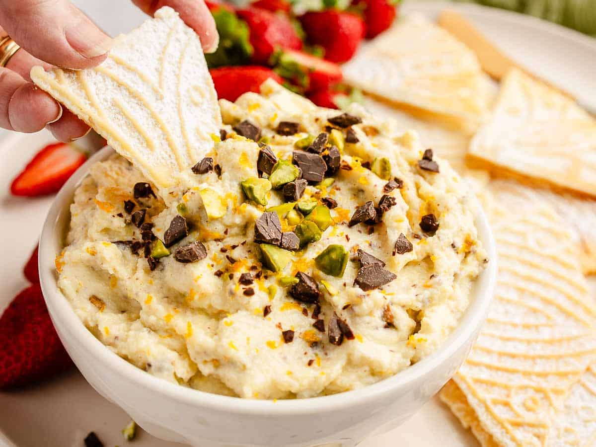 Close up of a creamy ricotta dip topped with chocolate chunks and pistachios with pizzelle being used to dip.