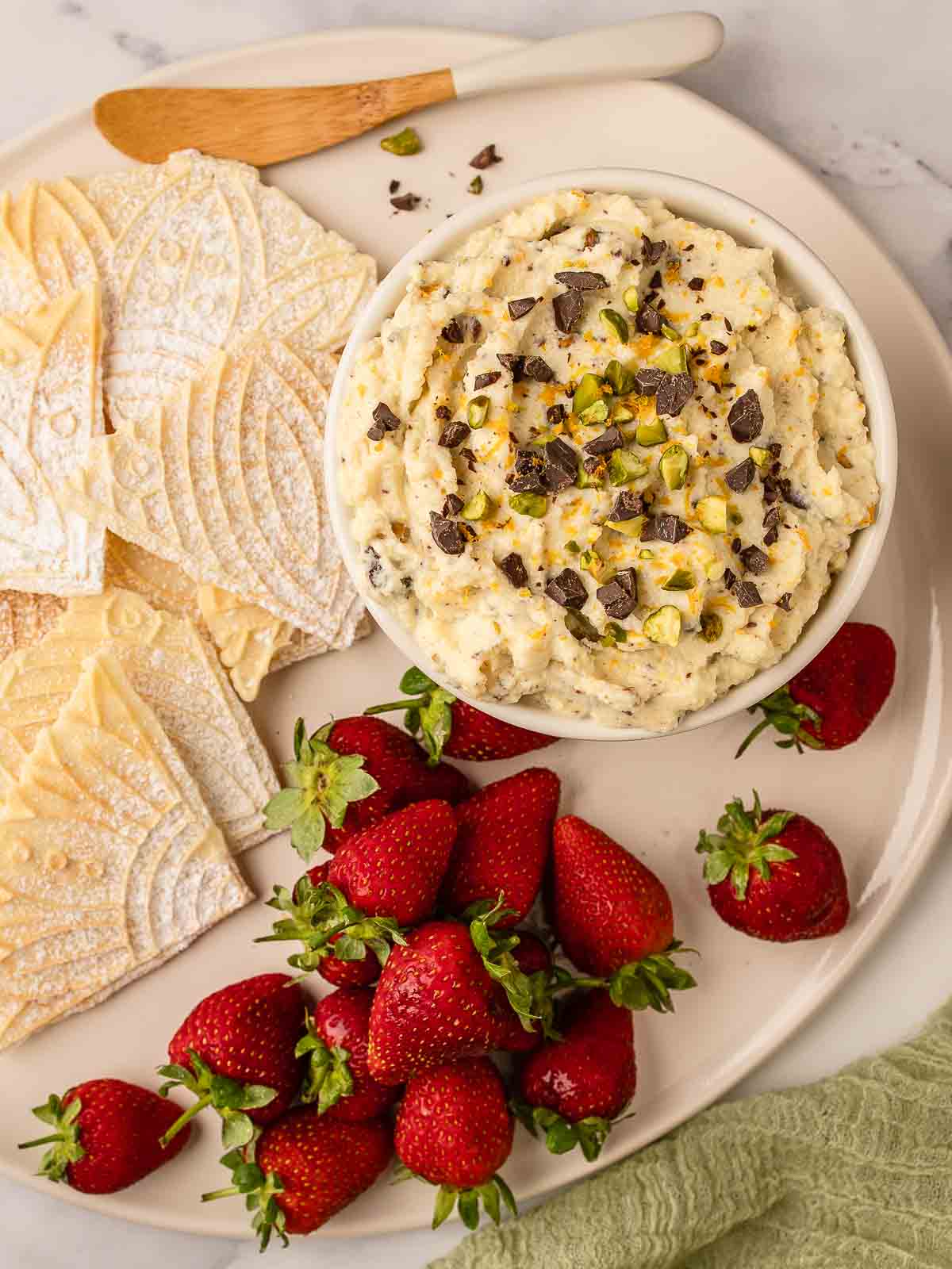 Overhead view of a creamy ricotta dip topped with chopped chocolate and pistachio nuts served with pizzelle and strawberries.