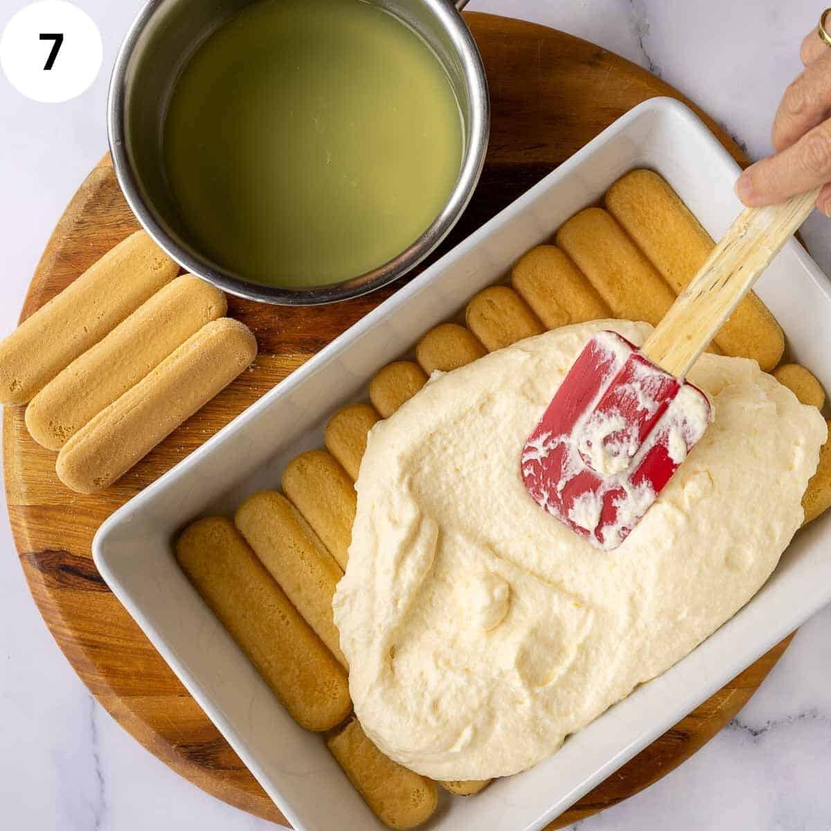 Spreading creamy mixture over savoiardi cookies in a rectangle dish.