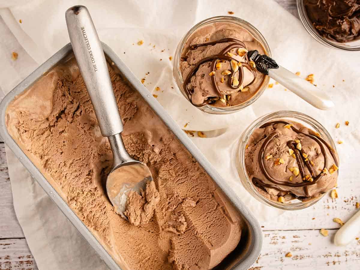 Overhead view of chocolate ice cream in a loaf pan with a ice cream scoop and two bowls of served ice cream on the side.