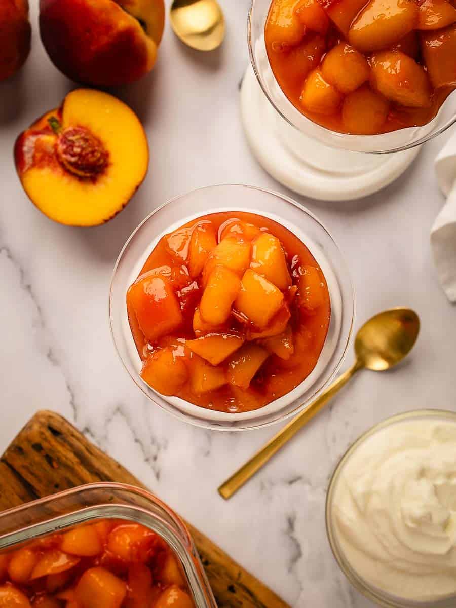 Overhead view of a compote of peaches in a glass with a halved peach and dishes of cooked peaches around.